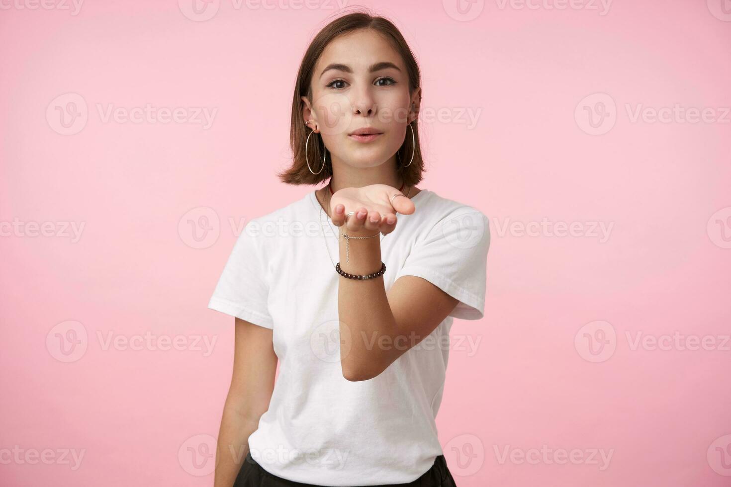 Horizontal shot of young brown-eyed brunette woman with bob haircut folding lips in air kiss and raising her hand while standing over pink background in casual wear photo