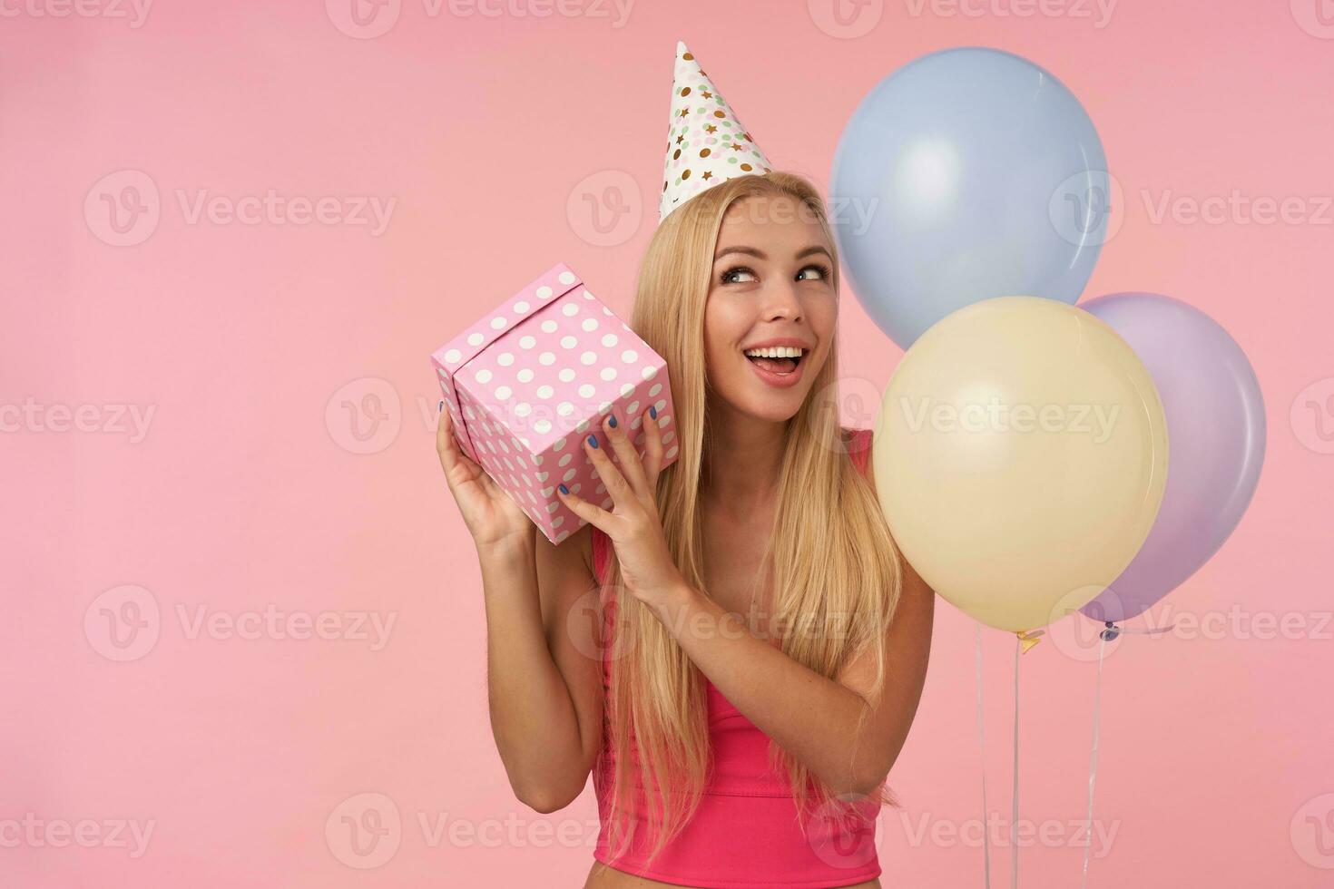 Happy young long haired blonde lady holding gift-wrapped box and wondering what is inside, rejoices nice party together with friends, standing over pink background and multicolored air balloons photo