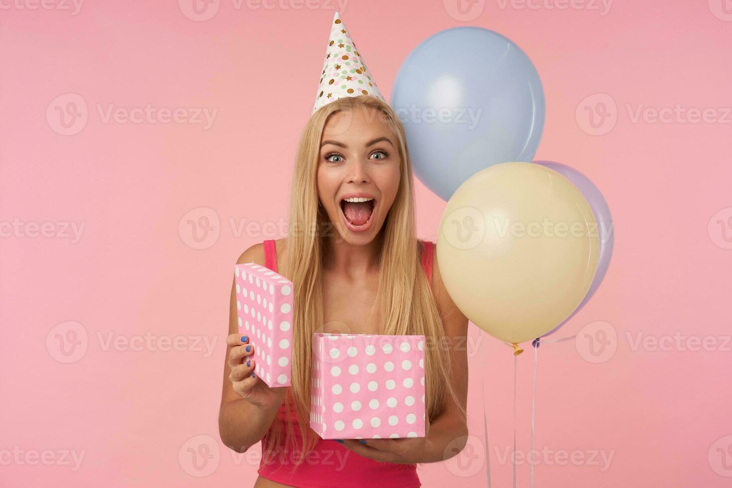 Overjoyed pretty young blonde woman lady with casual hairstyle showing happy reaction on getting awesome present, posing over pink background in birthday hat,looking at camera with wide cheerful smile photo