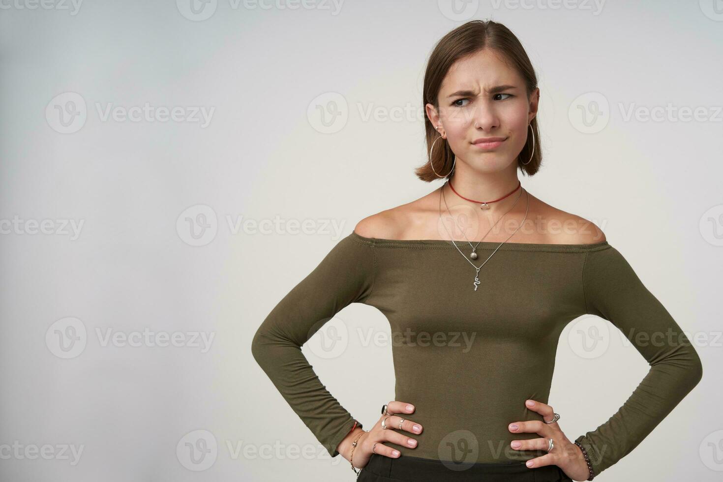 Studio shot of puzzled young pretty short haired brunette woman keeping hands on her waist while looking confusedly aside, posing over white background photo