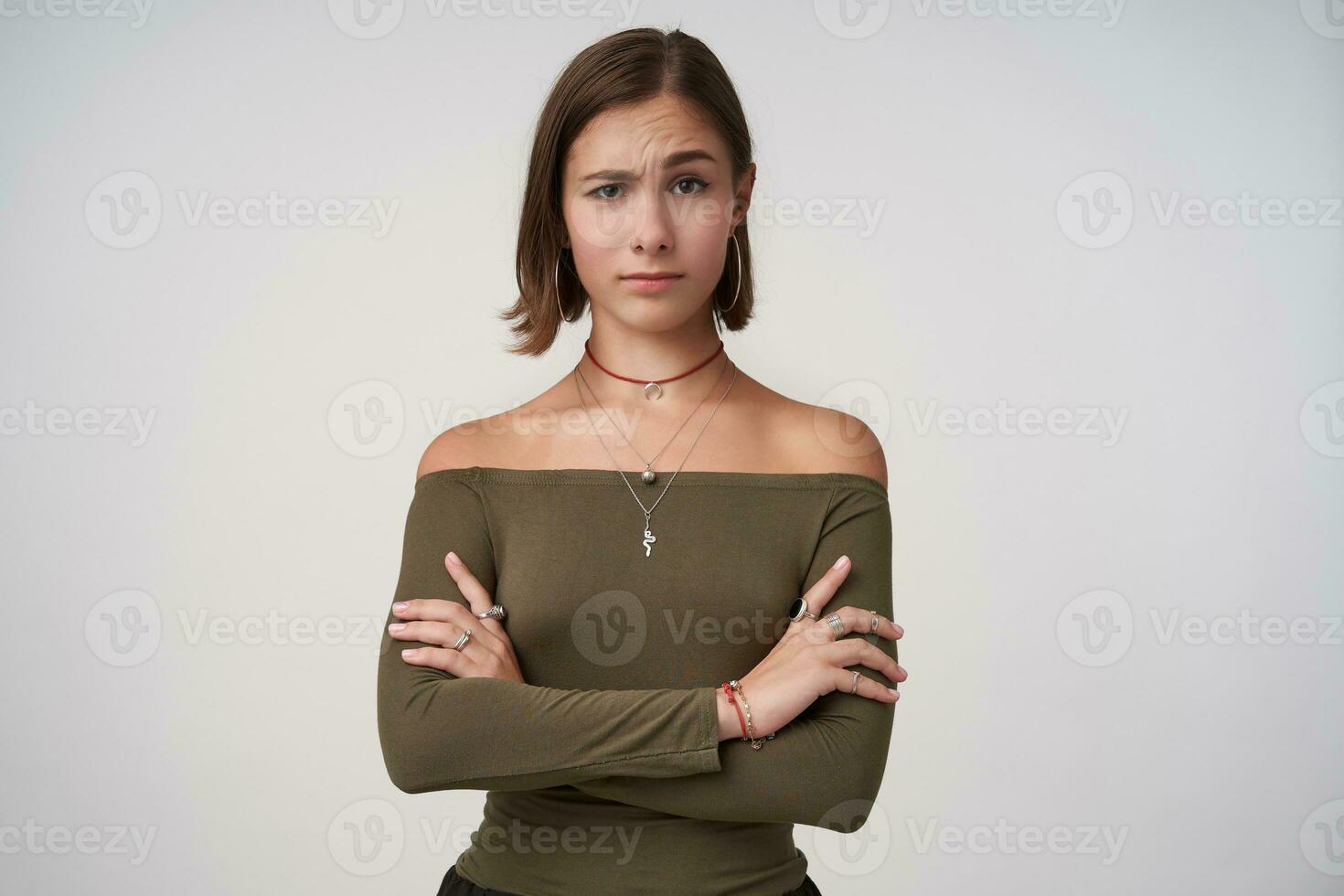 Indoor photo of young puzzled short haired brunette woman raising surprisedly eyebrow while looking at camera and folding hands on chest, isolated over white background