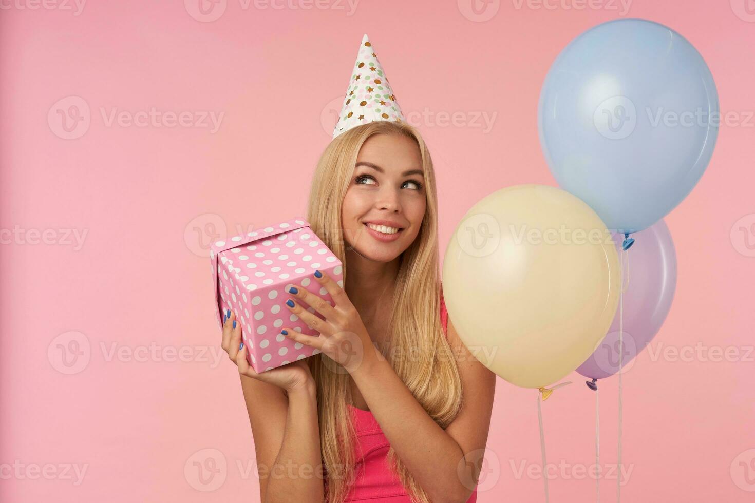 Portrait of cheerful young blonde female in pink top and birthday cap posing in multicolored air balloons, looking aside happily and keeping present box in hands, standing over pink background photo