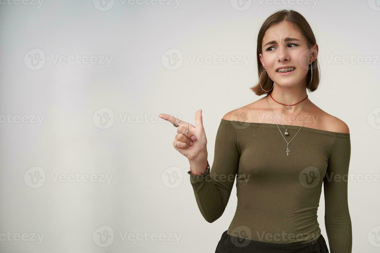 Indoor shot of displeased young short haired brunette woman frowning her face while showing aside with index finger, standing against white background photo