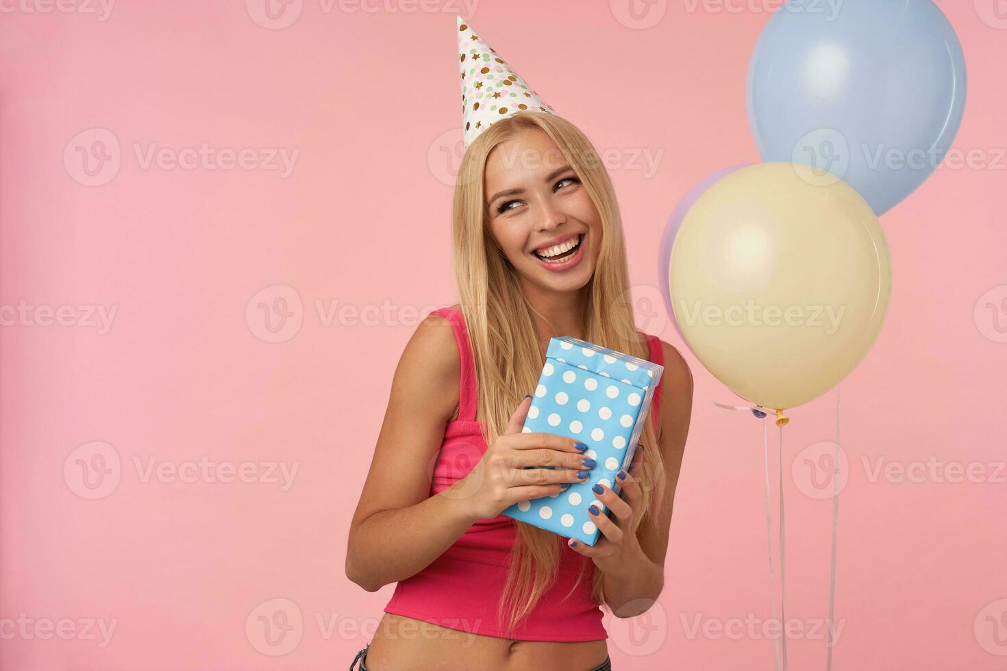 Pleasant looking cheerful young blonde female wearing pink top and holiday cone hat while standing over pink background, rejoicing while posing in multicolored air balloons photo