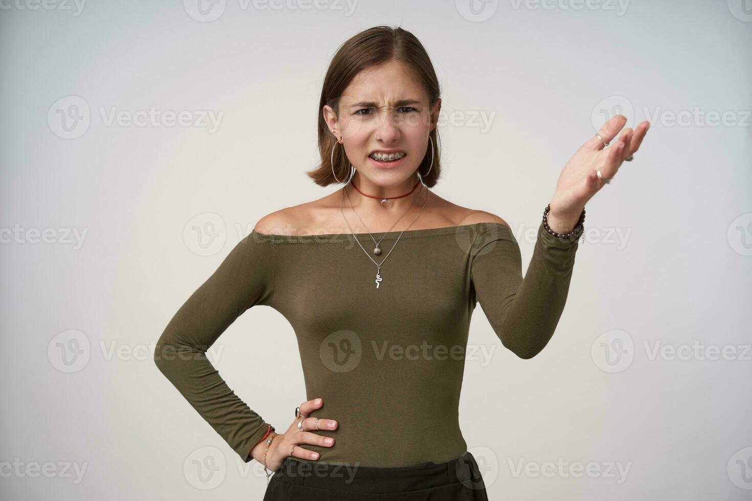 Disgruntled young short haired brunette woman with casual hairstyle keeping her hand raised while looking discontentedly at camera, isolated over white background photo