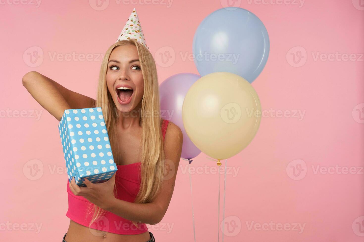 Joyful young blonde female with long hair posing over pink background while unpacking gifts, being excited and surprised to get birthday presents. People, entertainment and holiday attributes photo
