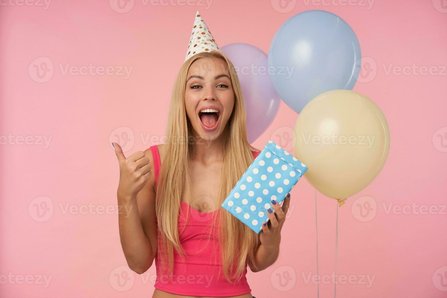 Joyful young blonde female with casual hairstyle in pink top and birthday cone cap standing over pink background and bunch of multicolored helium balloons, looking at camera and raising thumb happily photo