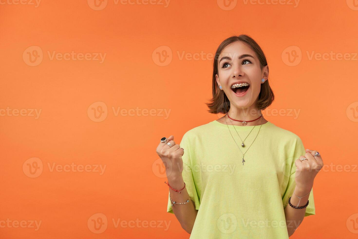 Young pretty woman with short brunette hair smiling. Very excited girl look to left upper corner at the copy space over orange background. Wearing green t-shirt, necklace, bracelets and rings photo