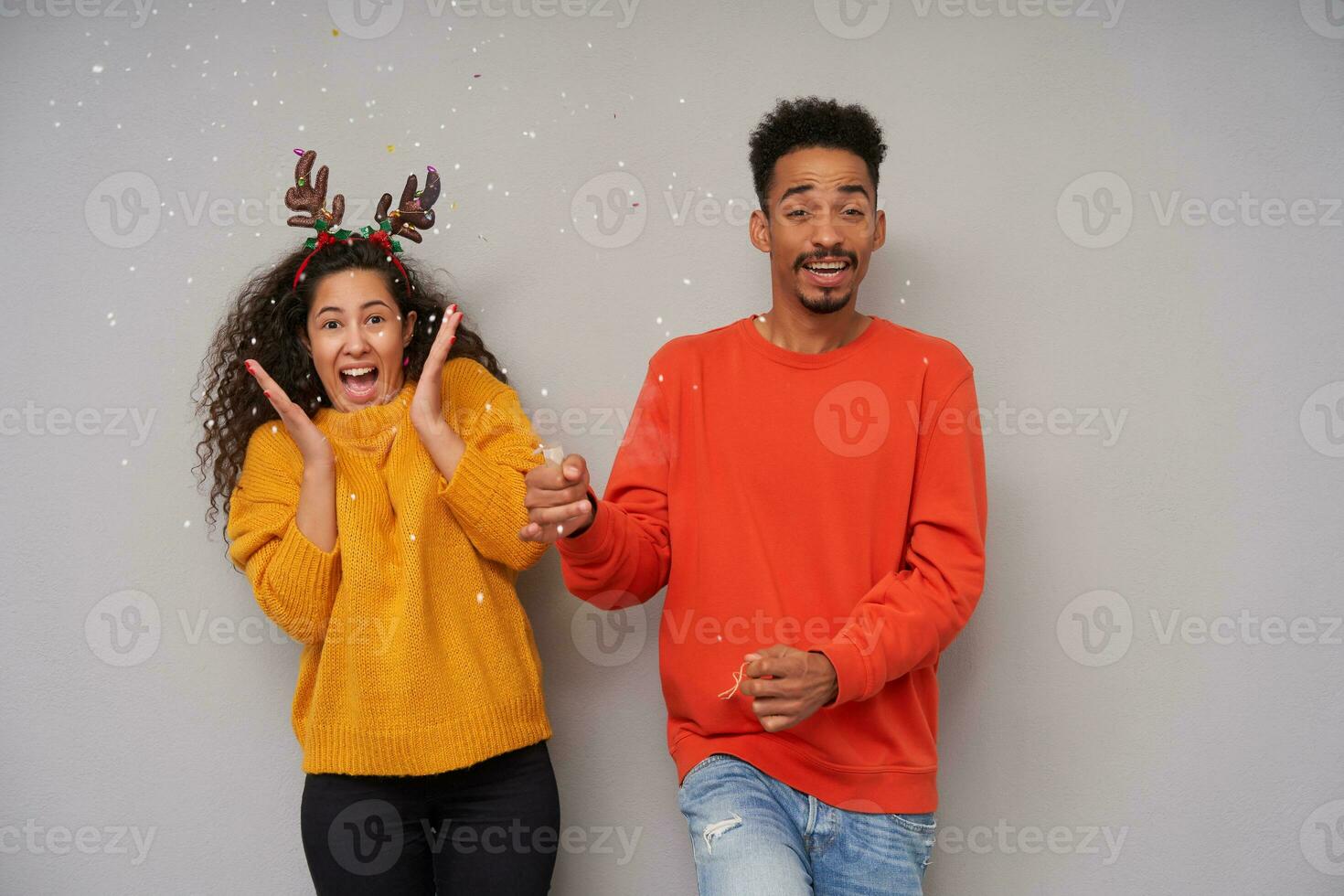 Indoor photo of joyful young dark skinned curly pair making confetti with clapper while celebrating new year, wearing cosy woolen colourful sweaters and holiday hoop over grey background