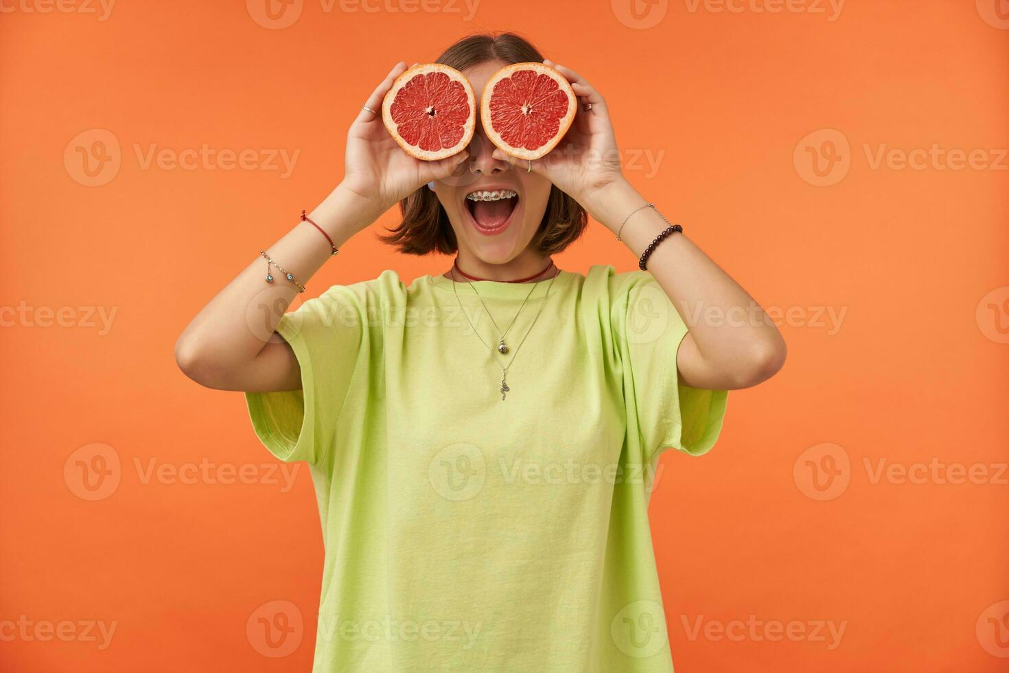 Female student, young lady with short brunette hair holding grapefruit over her eyes. Surprised looking. Standing over orange background. Wearing green t-shirt, teeth braces and bracelets photo