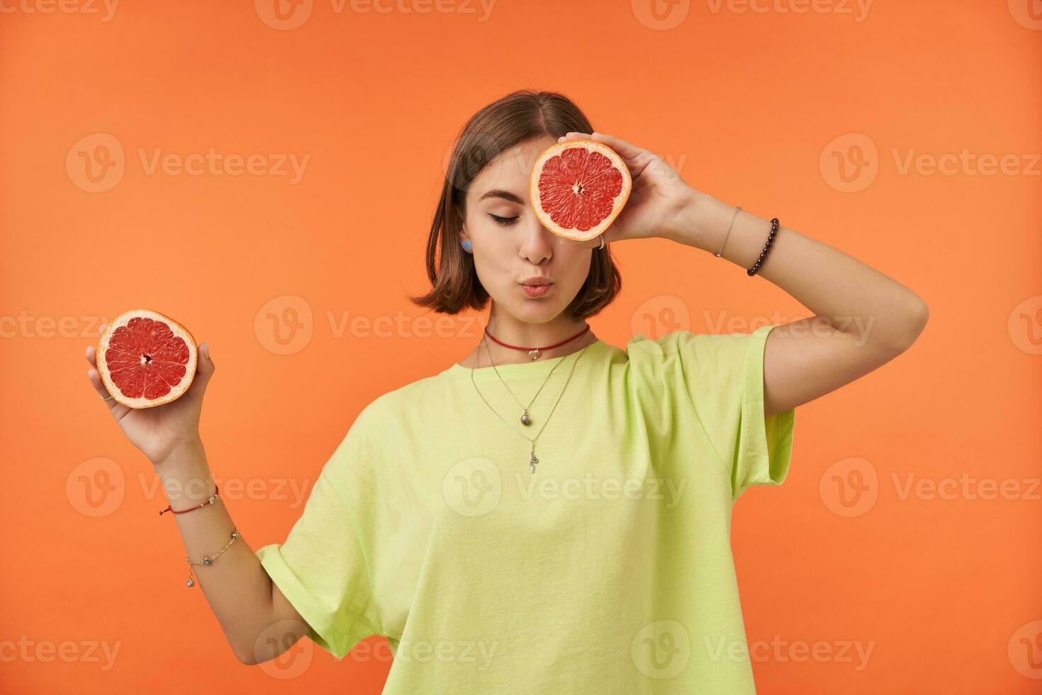 Female student, young lady with short brunette hair, dreaming with fruits. Holding grapefruit over her eye, cover one eye. Standing over orange background. Wearing green t-shirt, braces and bracelets photo