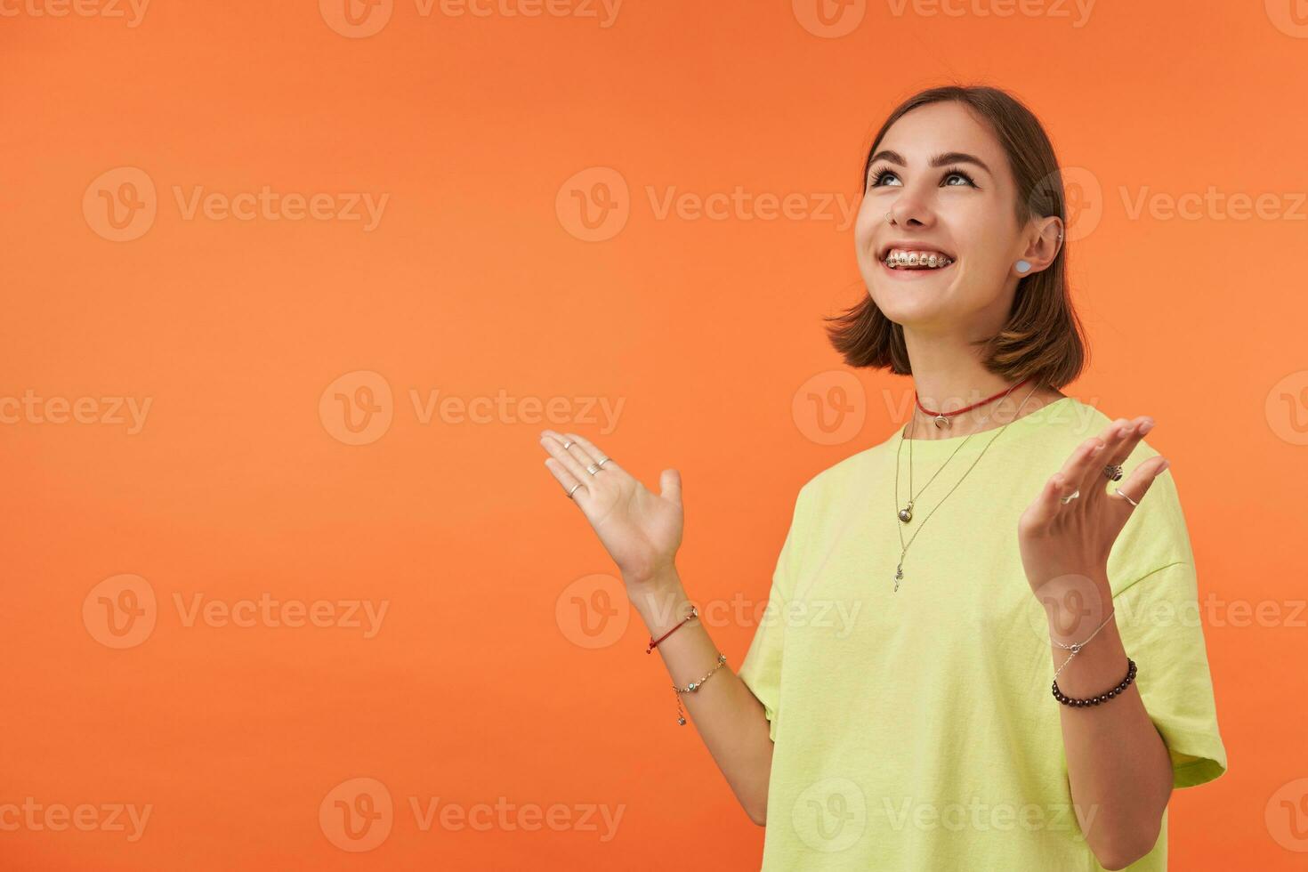 Teenage girl, cheerful and happy looking woman with brunette short hair. Happy to share feelings, feels lucky. Watch to the left and up at the copy space over orange background. Wearing green t-shirt photo