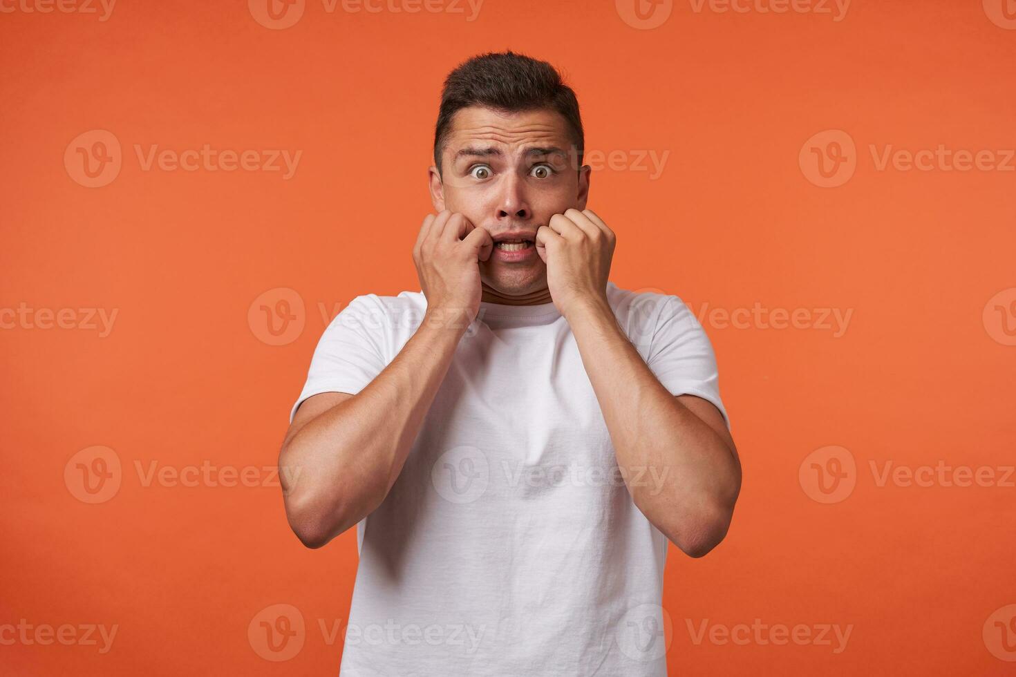 Studio photo of young pretty brown haired male holding raised hands on his cheeks while looking scaredly at camera, standing against orange background