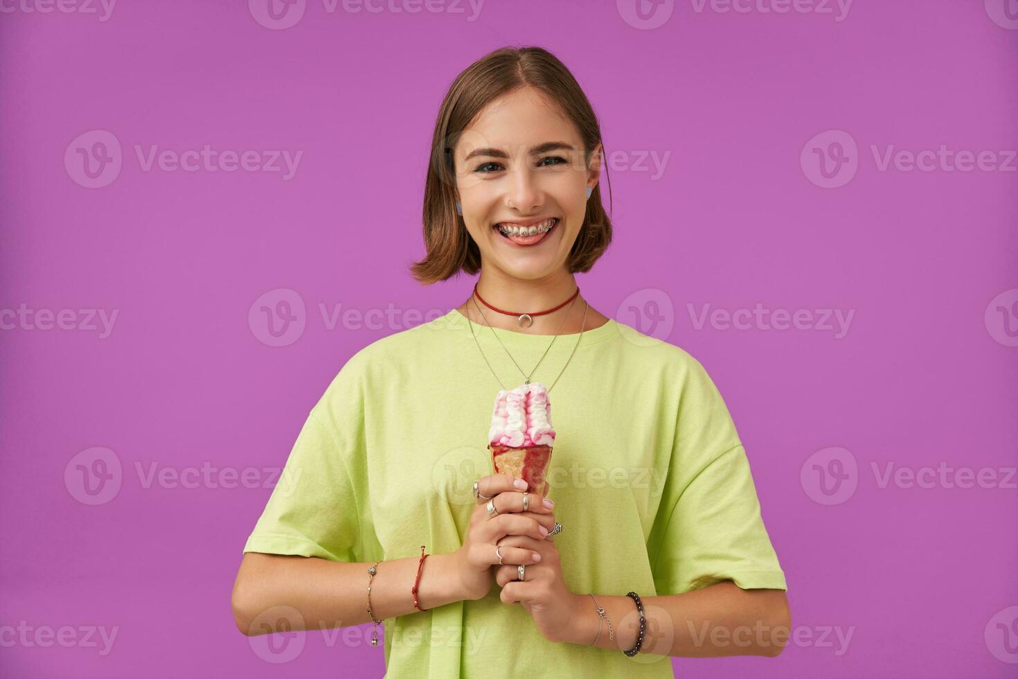 hembra alumno, joven dama con hielo crema en ambos manos, demostración su lengua y sonriente a el cámara, terminado púrpura antecedentes. vistiendo verde camiseta, esposas, anillos y collares foto