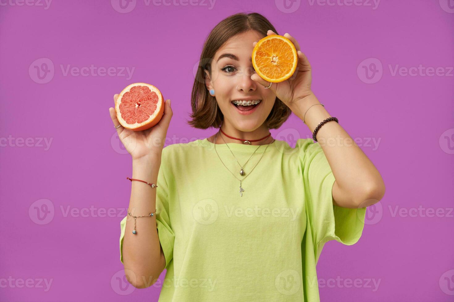 Female student, young surprised lady with short brunette hair. Holding orange over her eye, cover one eye. Standing over purple background. Wearing green t-shirt, necklace, braces and bracelets photo