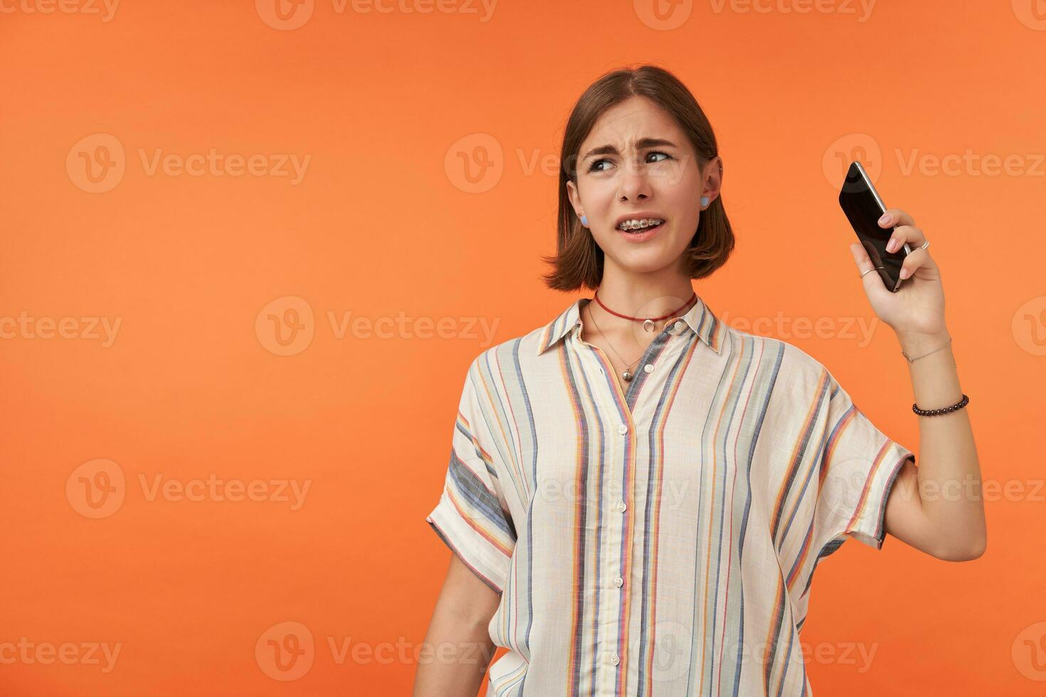 adulto niña mirando infeliz y colgando arriba un teléfono. diferente a llamar. estudiante recoger arriba un teléfono, vistiendo a rayas camisa, dientes tirantes y esposas. acecho a el izquierda a Copiar espacio terminado naranja antecedentes foto