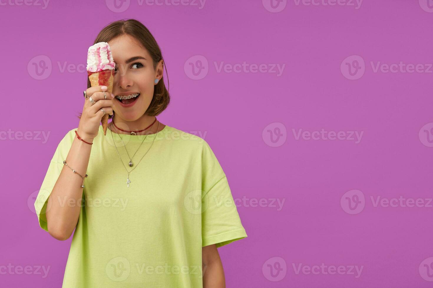 Teenage girl, cheerful and happy, with brunette short hair. Holding ice cream over her eye and looking to the right at the copy space over purple background. Wearing green t-shirt, rings and necklace photo