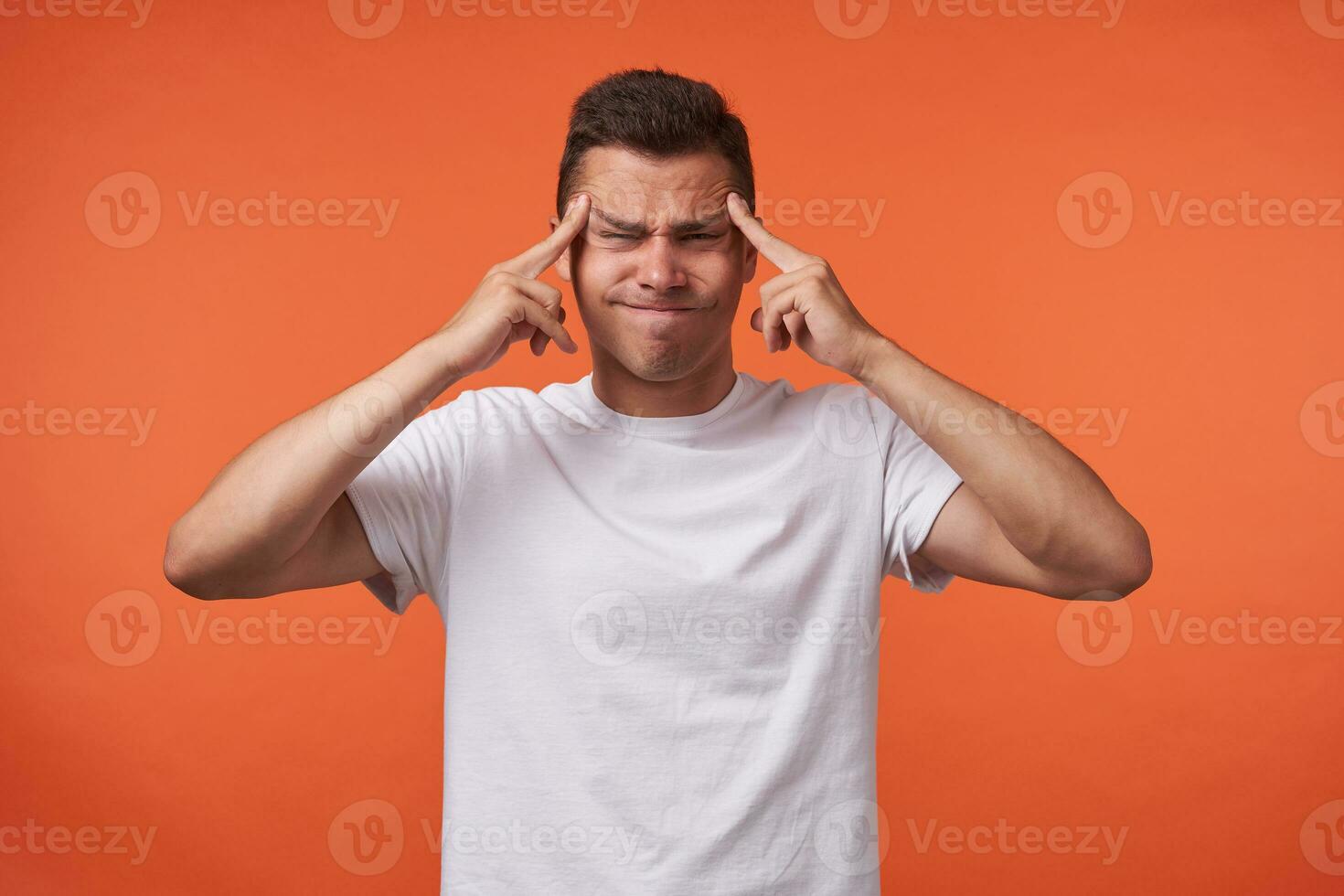 Unhappy young handsome brown haired man with short haircut keeping fingers on temples and frowning face while standing over orange background in white basic t-shirt photo