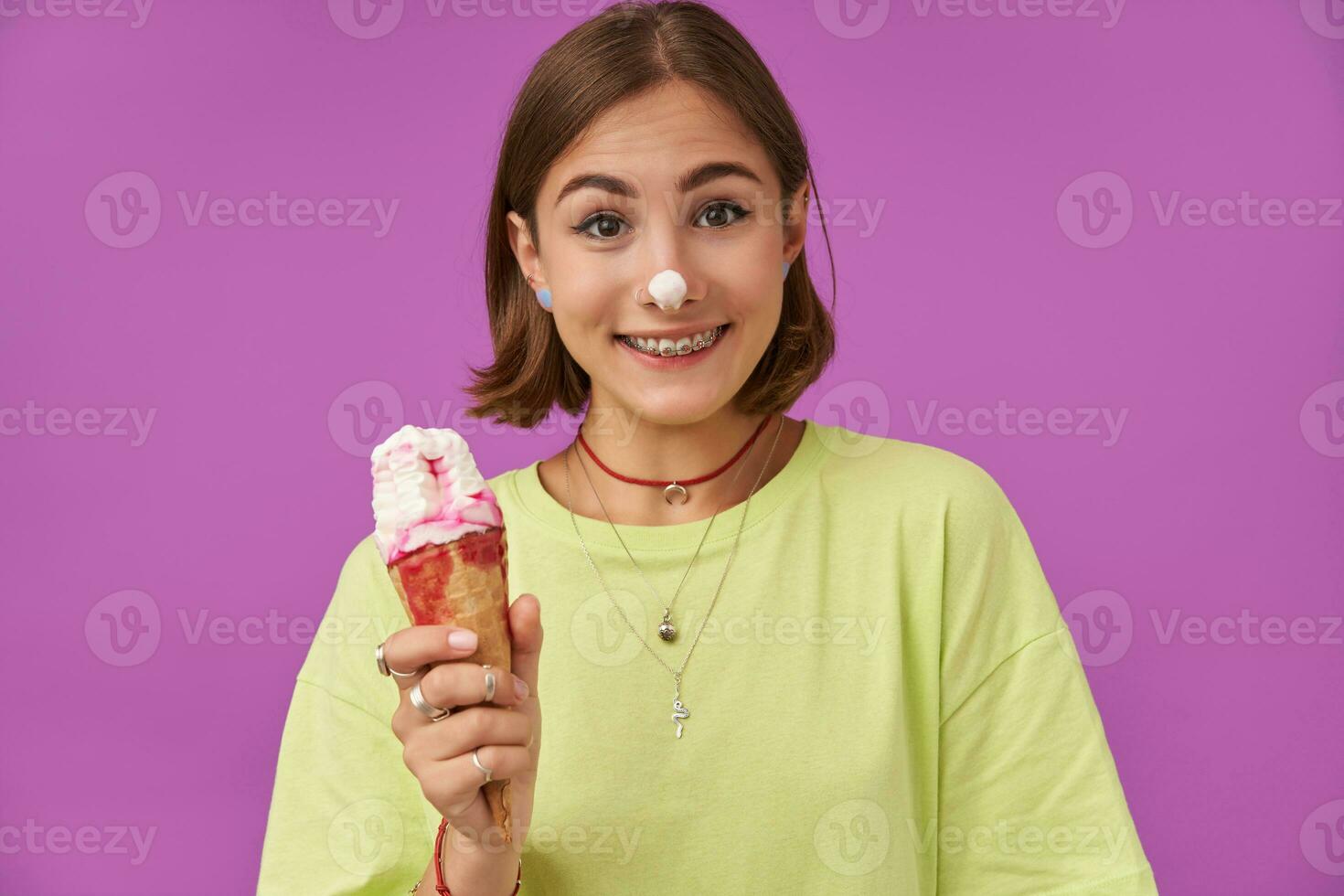 Teenage girl, cheerful and happy, with brunette short hair, ice cream on a nose. Holding an ice cream and smile at the camera, over purple background. Wearing green t-shirt, teeth braces and bracelets photo