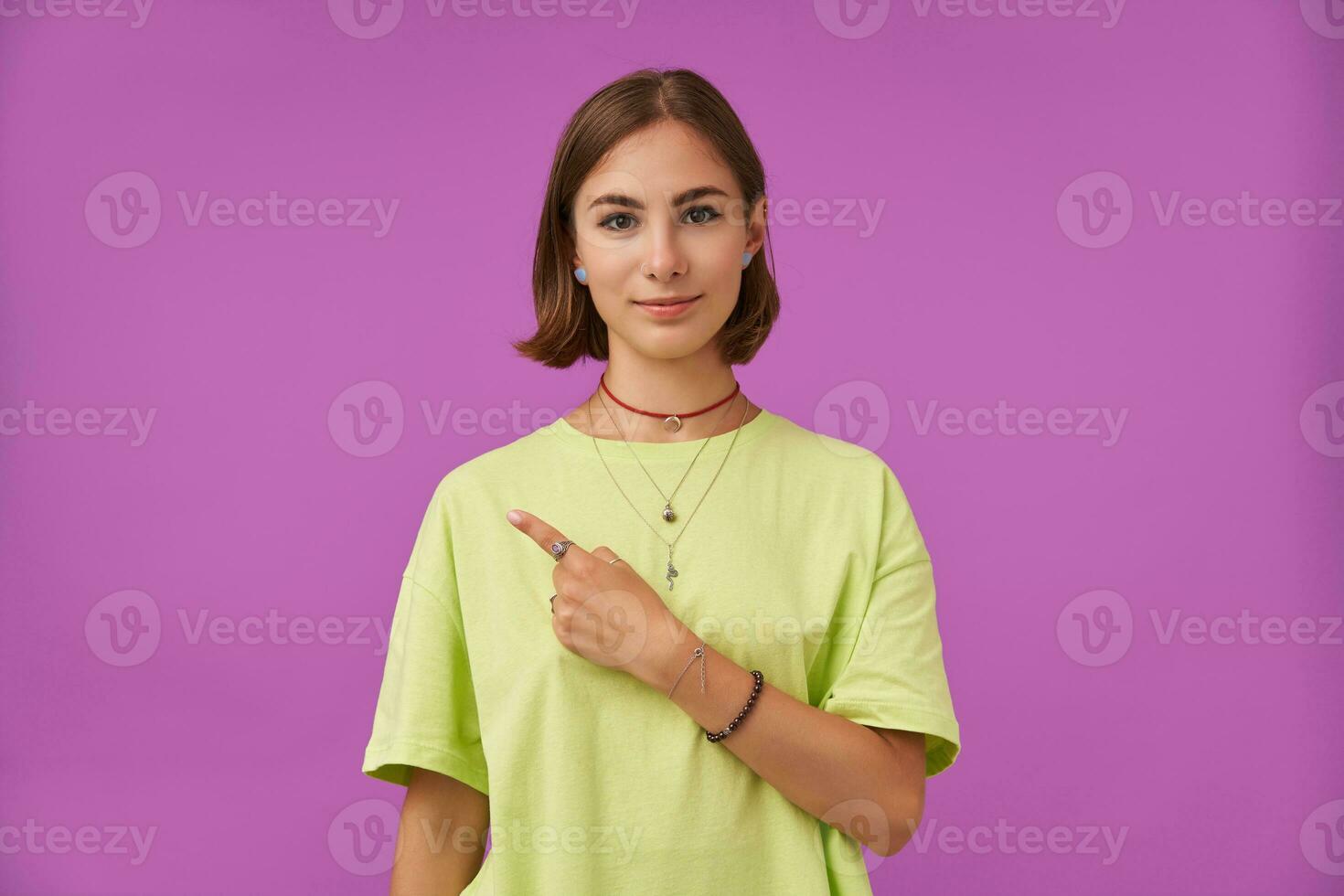 Female student, young lady with brunette short hair, pointing her finger to the left at the copy space over purple background. Showing a sign. Wearing green t-shirt, bracelets and rings photo