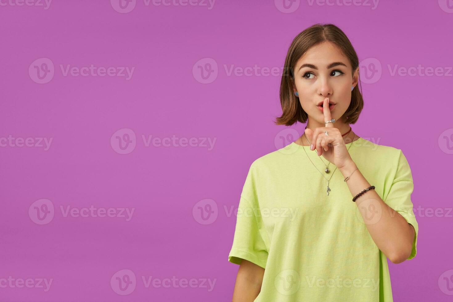 Portrait of attractive, nice looking girl. Touching lips with finger, showing a silence sign. Watching to the left at the copy space over purple background. Wearing green t-shirt, bracelets and rings photo