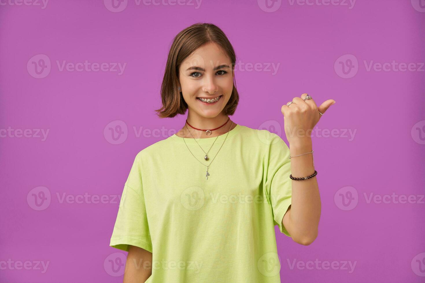 Portrait of attractive, nice looking girl. Smiling and pointing a thumb to the right at the copy space over purple background. Wearing green t-shirt, teeth braces, necklace and bracelets photo