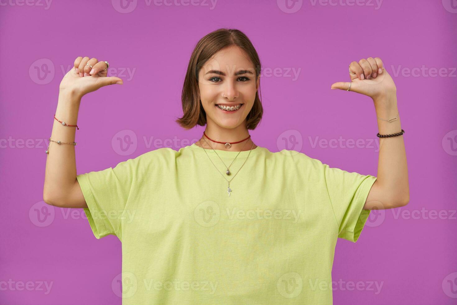 Teenage girl, cheerful and happy, with brunette short hair. Pointing thumbs at herself and smile. Standing over purple background. Wearing green t-shirt, teeth braces, bracelets and rings photo