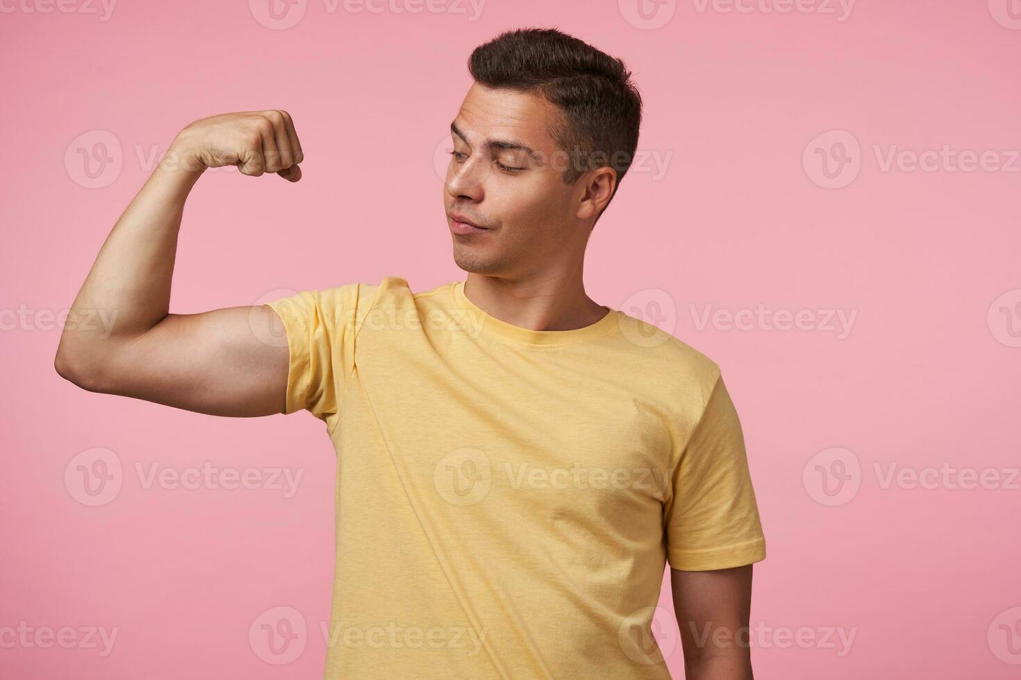 Studio photo of young handsome brown haired man looking proudly at his hand while demonstrating his strong biceps, standing over pink background in casual wear