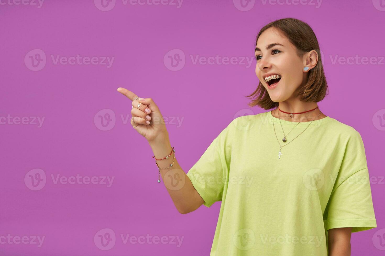 Female student, young lady, laughing and pointing her finger to the left at the copy space over purple background. Showing a sign. Wearing green t-shirt, teeth braces, bracelets and rings photo