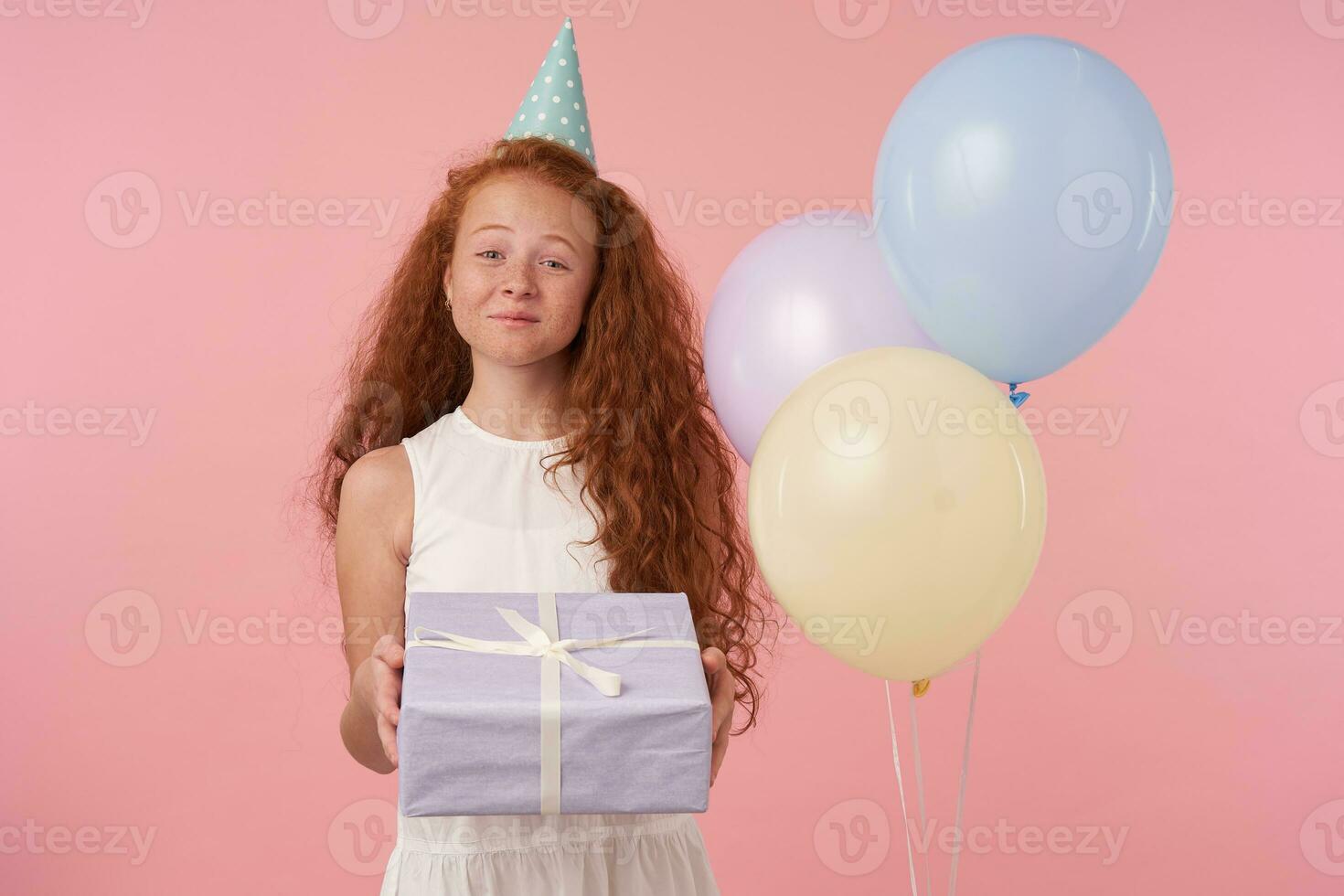 Photo of cute little girl with long foxy hair being amused by guests, smiles gladfully, isolated over pink studio background with air balloons, holding birthday presents and looking to camera happily