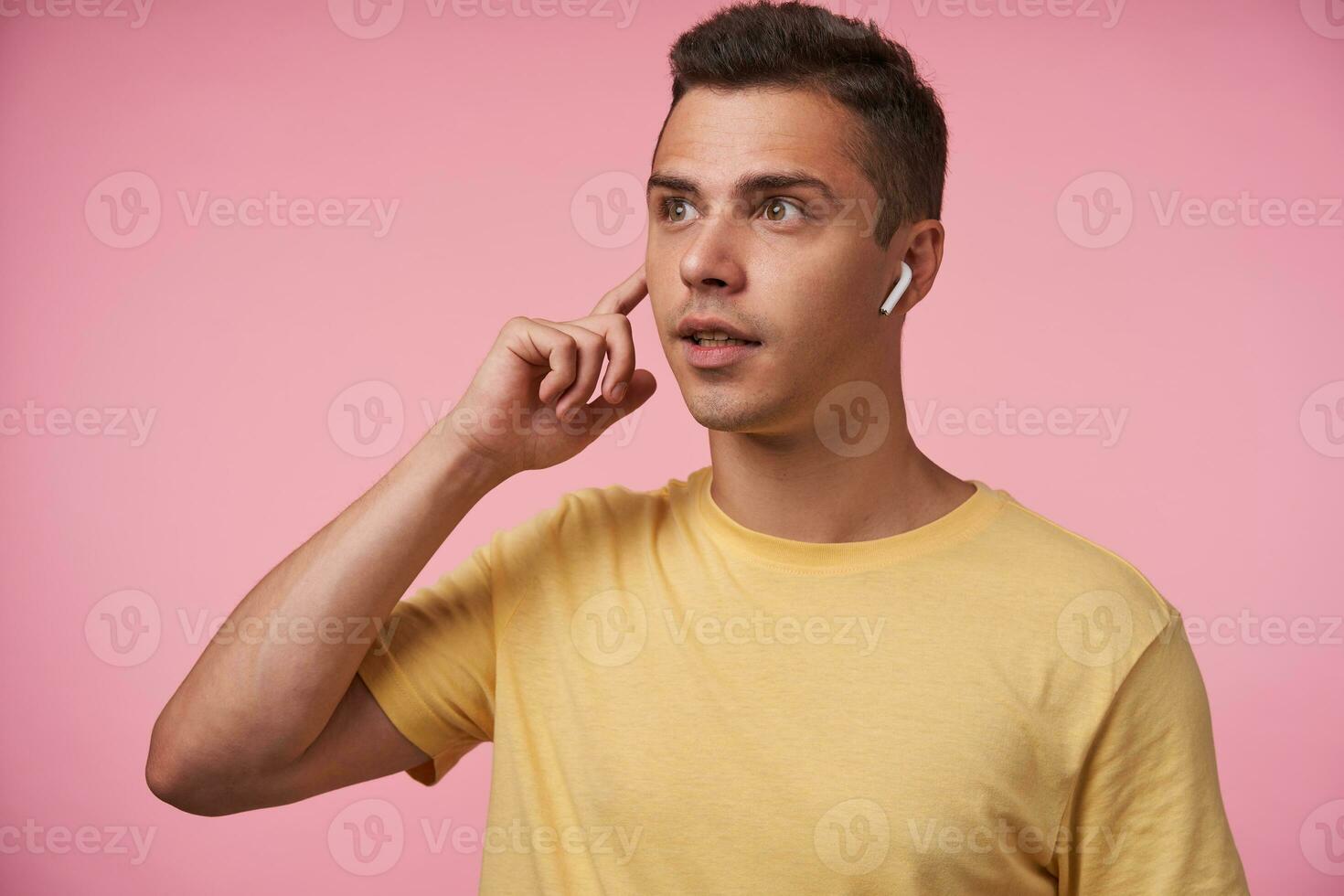 desconcertado joven bonito de ojos marrones morena chico vestido en amarillo camiseta acuerdo elevado dedo índice en auricular mientras mirando sorprendentemente adelante, aislado terminado rosado antecedentes foto