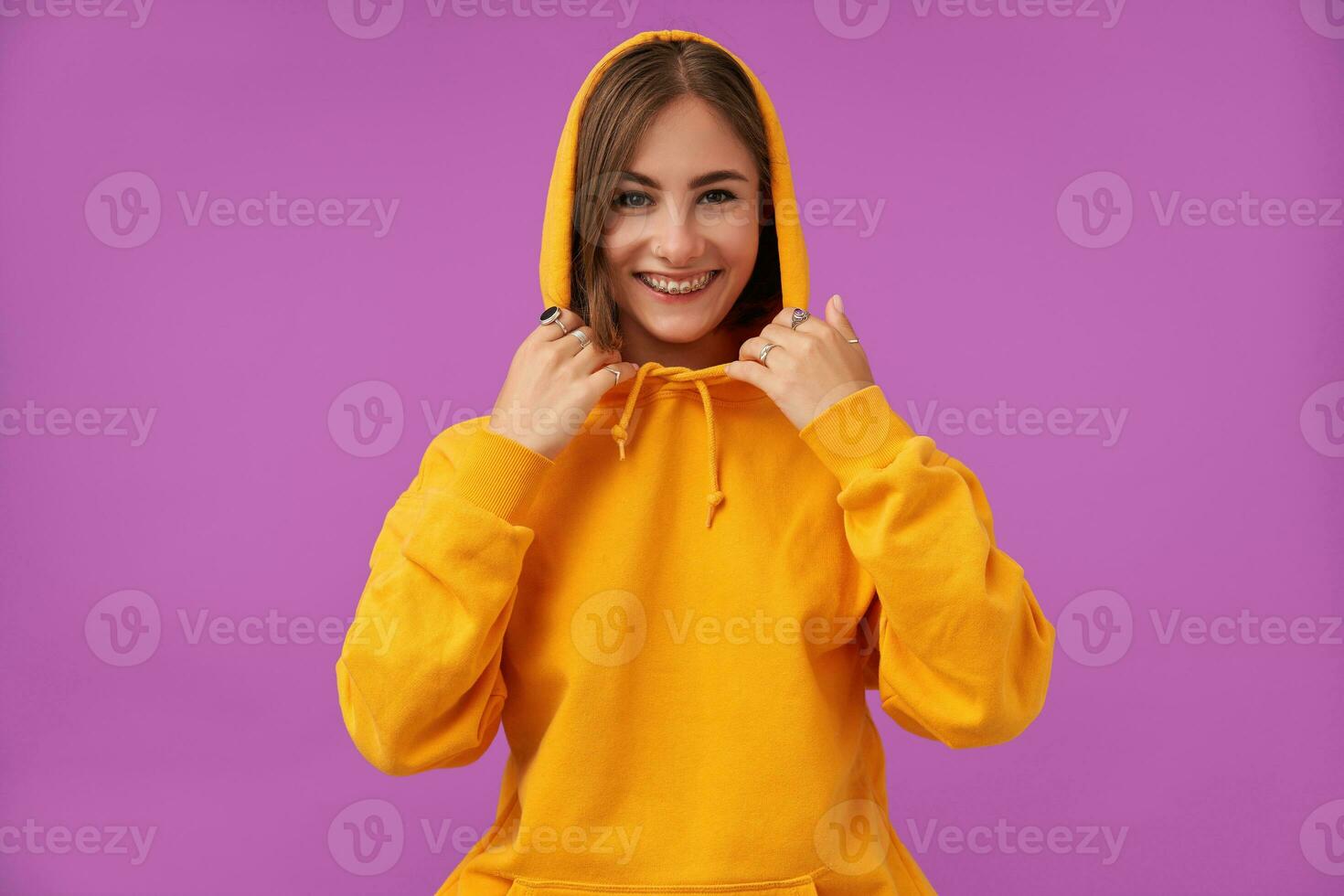 Portrait of attractive, nice looking girl with short brunette hair. Smiling at he camera and touching hood with hands, over purple background. Wearing orange hoodie, rings and teeth braces photo