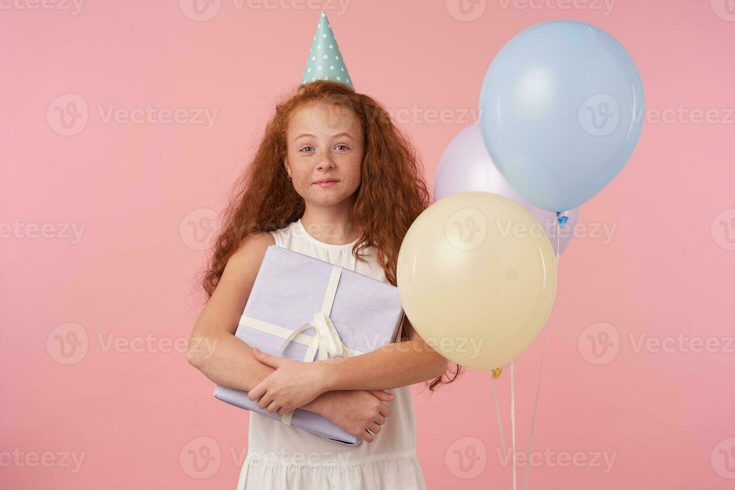 Portrait of lovely little curly girl with long foxy hair standing over pink background in white elegant dress, looking in camera with sincere charming smile, being in high spirit photo