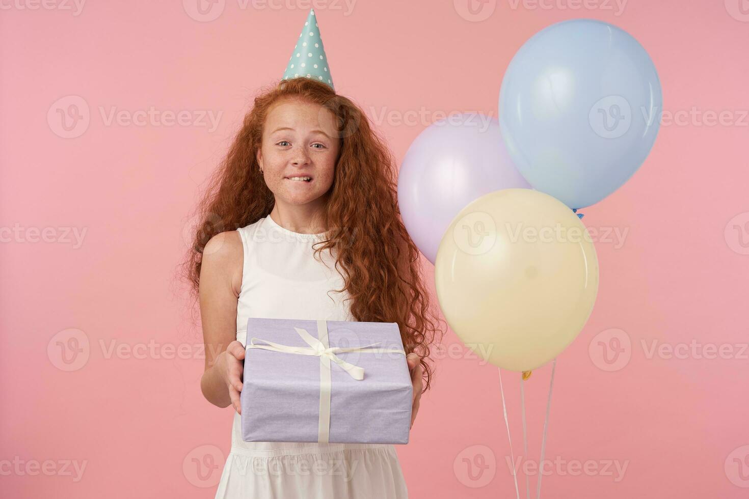 Overjoyed curly female kid with long foxy hair posing over pink background with gift-wrapped box and looking to camera cheerfully, wearing white elegant dress and birthday cap photo