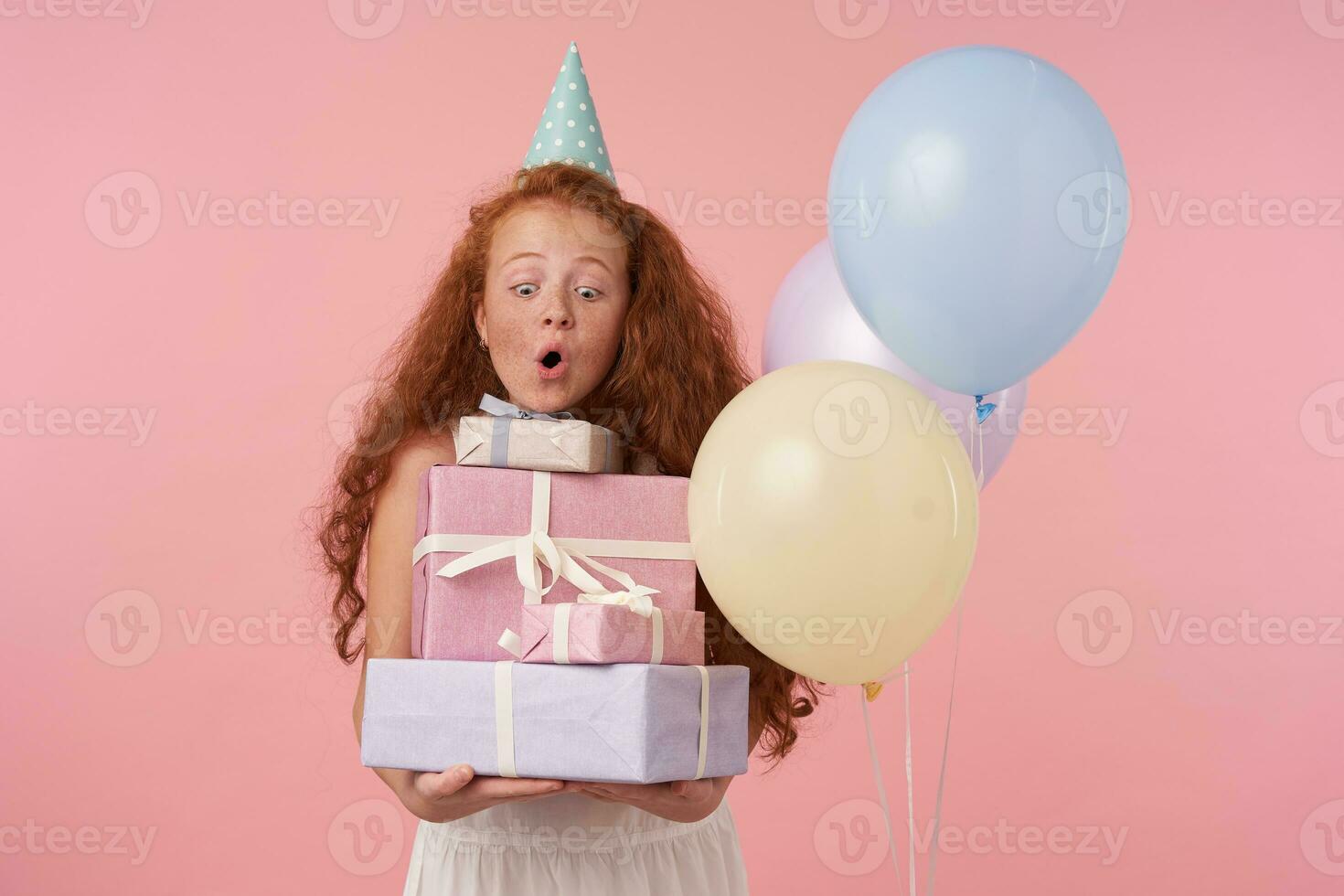 Indoor photo of redhead female kid in festive clothes and birthday cap posing over pink background with gift boxes in hands, being excited and surprised to get birthday presents