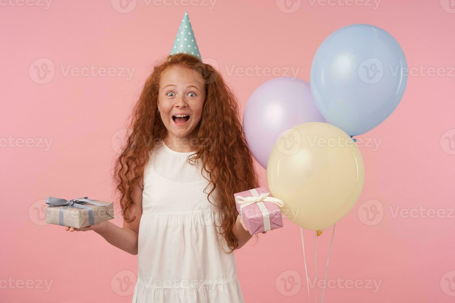 Joyous female curly kid with long foxy hair being excited and surprised to get birthday present, happily looking in camera with gift box in hands over pink background. Children and celebration concept photo