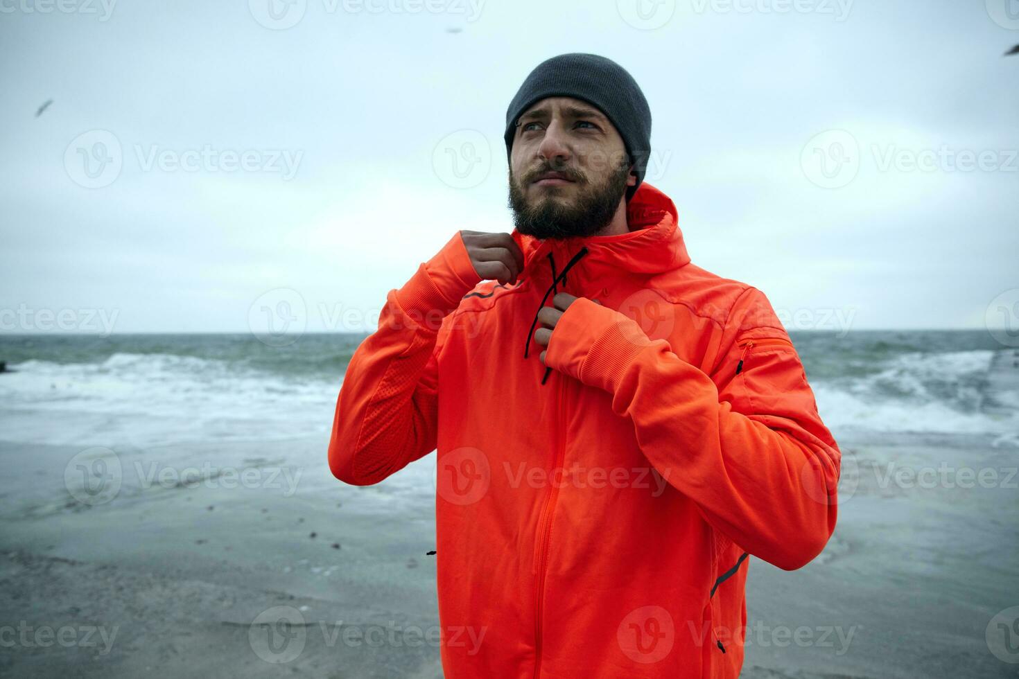 Sporty serious looking young dark haired bearded man with eyebrow piercing wearing black cap and winter sporty coat with hood, posing over coastline of sea on cold stormy weather photo