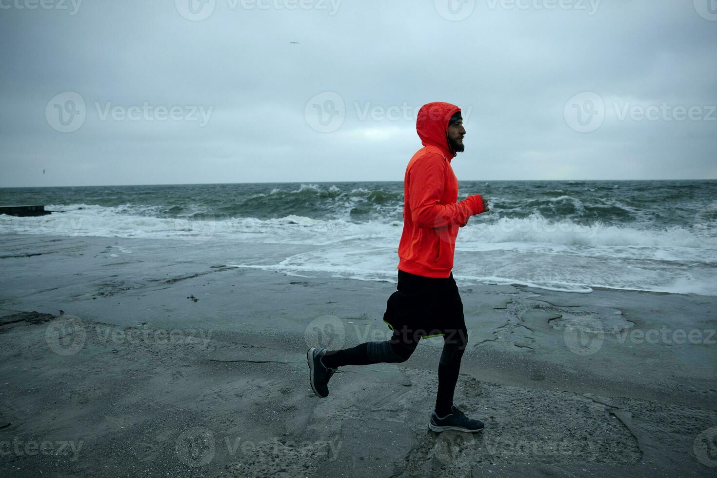 Image of young active bearded male doing morning exercises by sea, running along seafront on cold gloomy weather, wearing warm athletic clothes. Fitness male model photo
