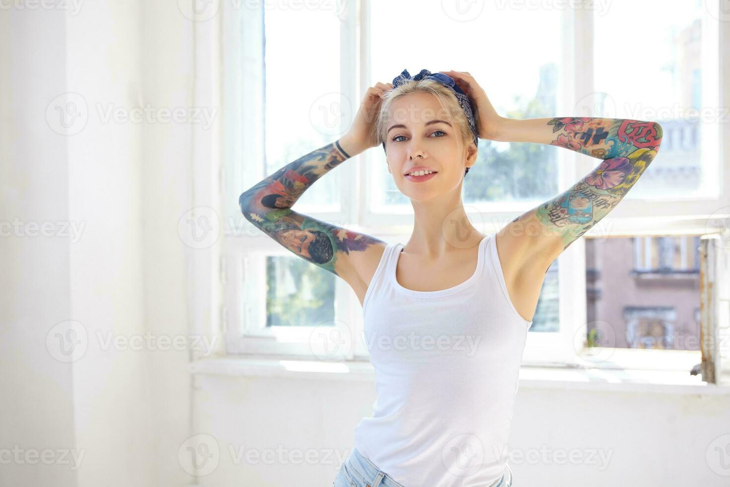 Sunny shot of young attractive tattooed blonde lady keeping raised hands on her head while looking at camera with pleasant smile, standing in front of big window photo