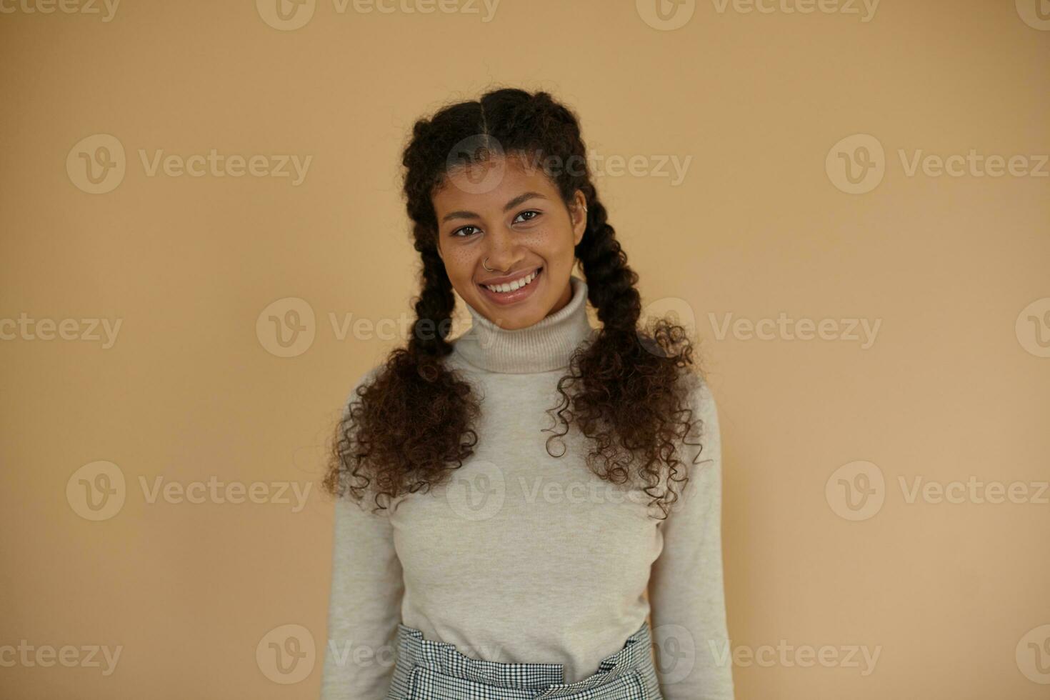 Indoor shot of pretty curly dark skinned brunette lady with braids looking on camera with charming smile while standing over beige background, keeping hands along body photo