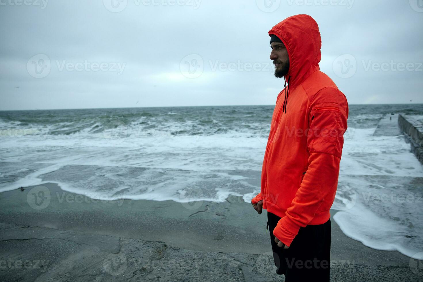 Horizontal shot of young dark haired bearded sportsman dressed in warm orange hoodie and black athletic trousers standing over seaside on grey early morning and looking thoughtfully ahead photo