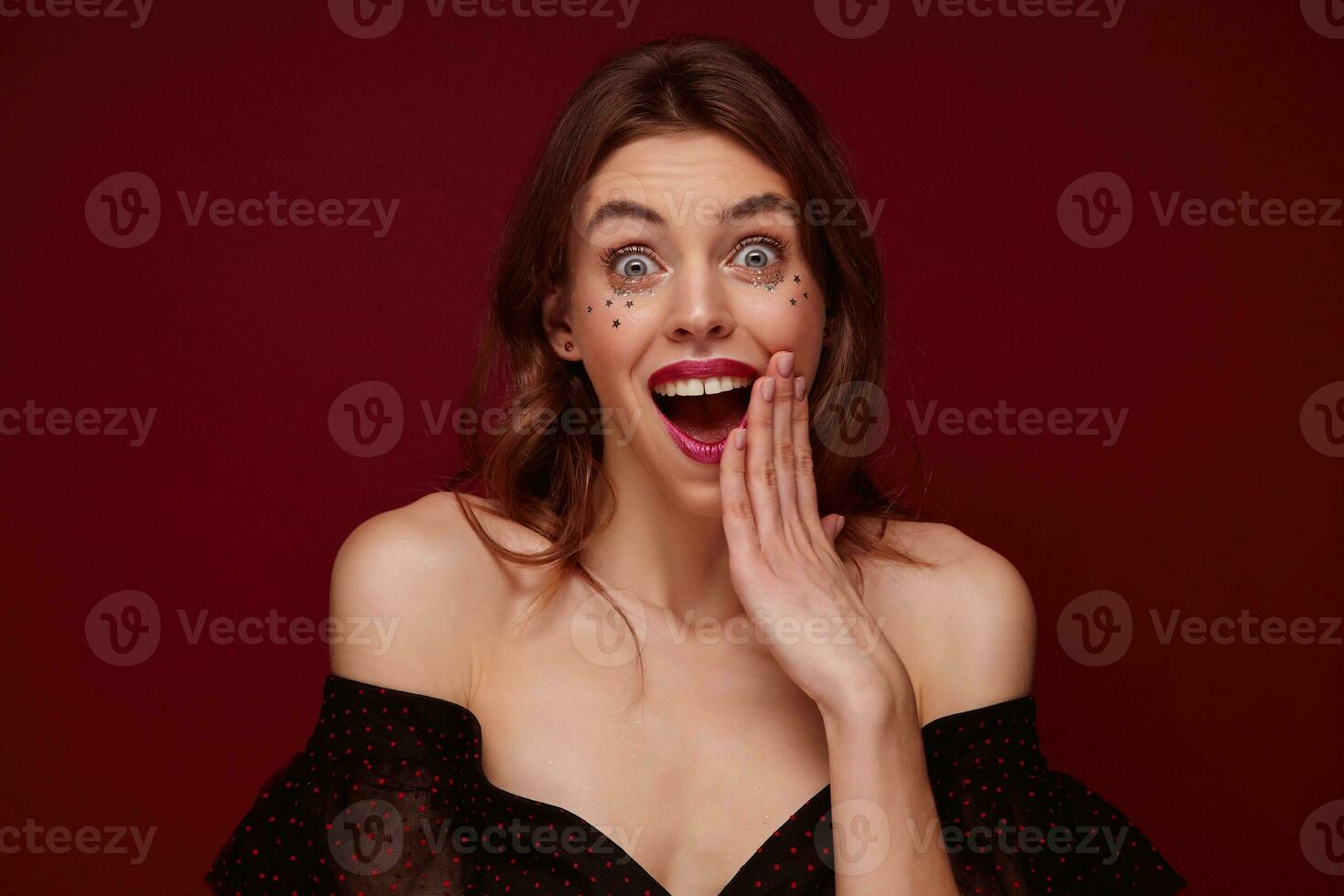 Surprised pretty young female with brown wavy hair looking at camera with wide eyes and mouth opened, keeping palm near her face, dressed in elegant black top with red dots over claret background photo