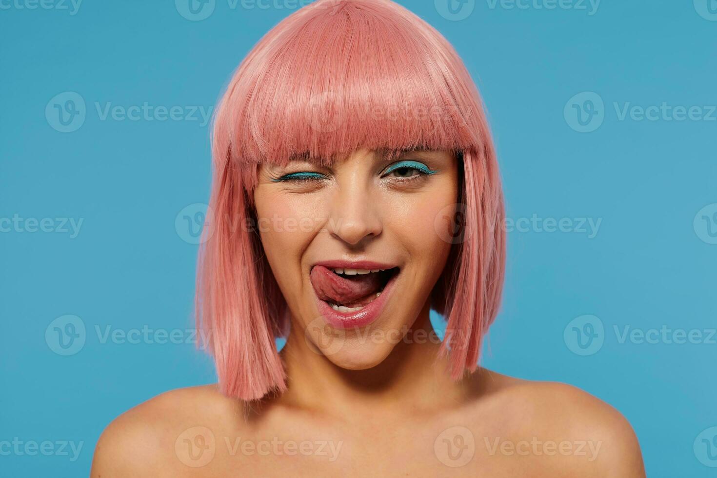 Coquettish young pretty woman with short pink hair giving wink while looking playfully at camera, sticking out her tongue while standing over blue background photo