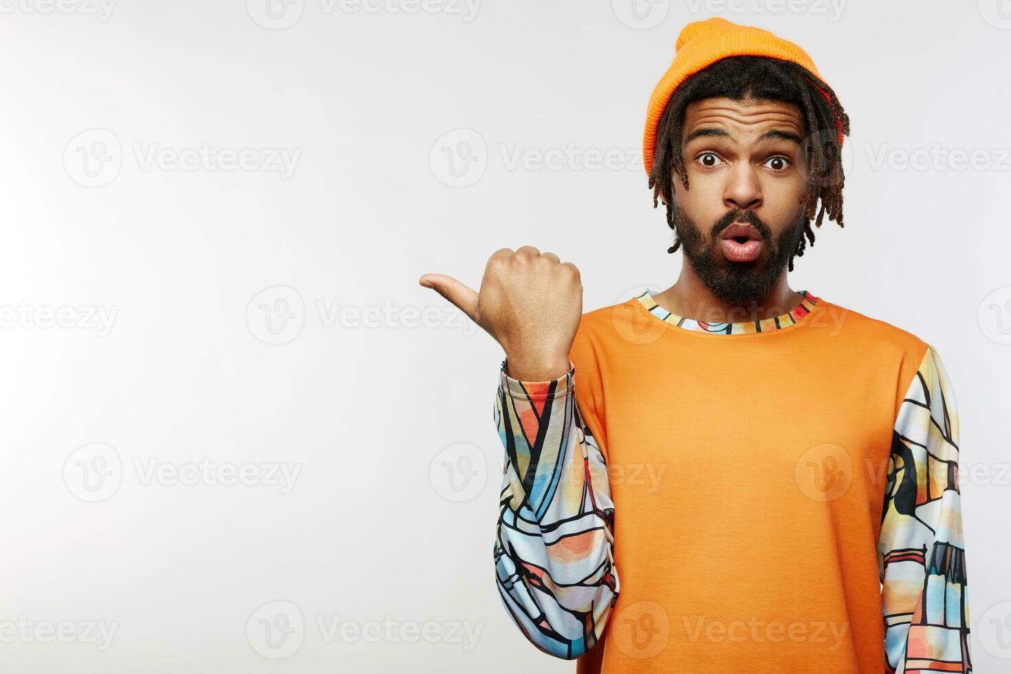 Surprised young dark haired male with dreadlocks wrinkling his forehead while looking amazedly at camera and pointing aside with raised hand, posing over white background photo