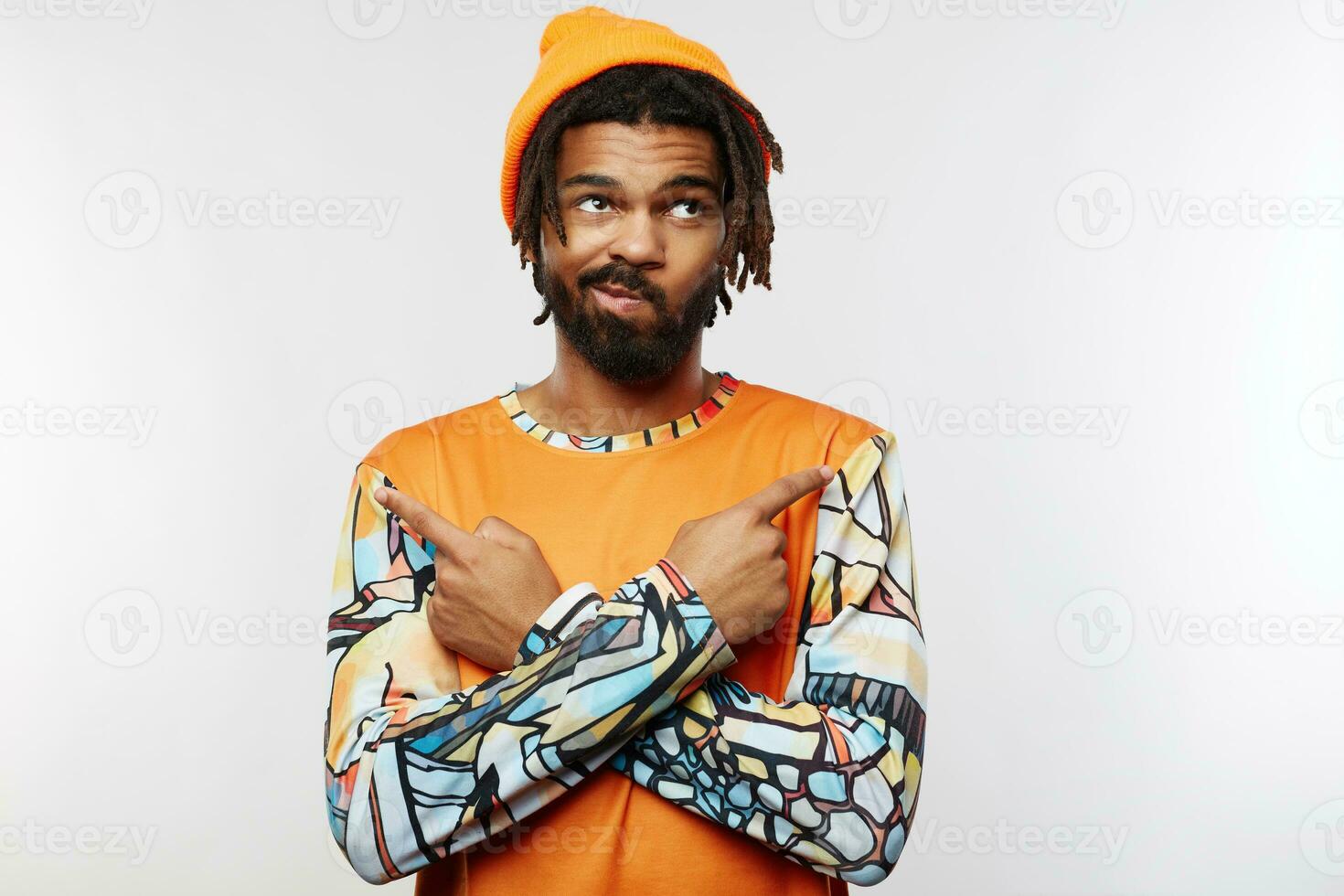 Displeased young handsome dark skinned guy with beard twisting his mouth while looking upwards and pointing in different sides with raised forefingers, isolated over white background photo
