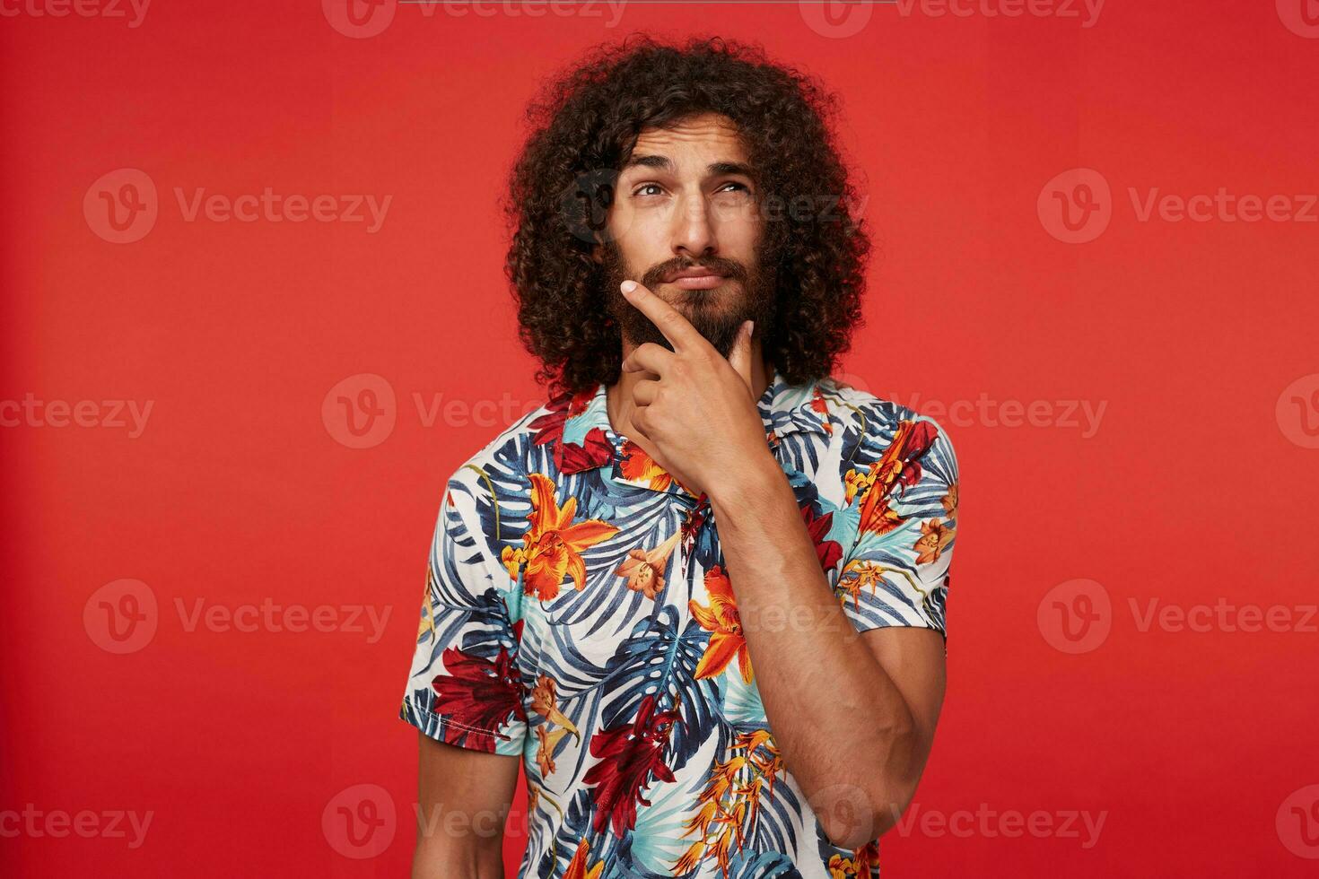 Pensive handsome young brunette curly male with beard holding his chin with raised hand and looking thoughtfully aside, squinting and wrinkling his forehead while standing over red background photo