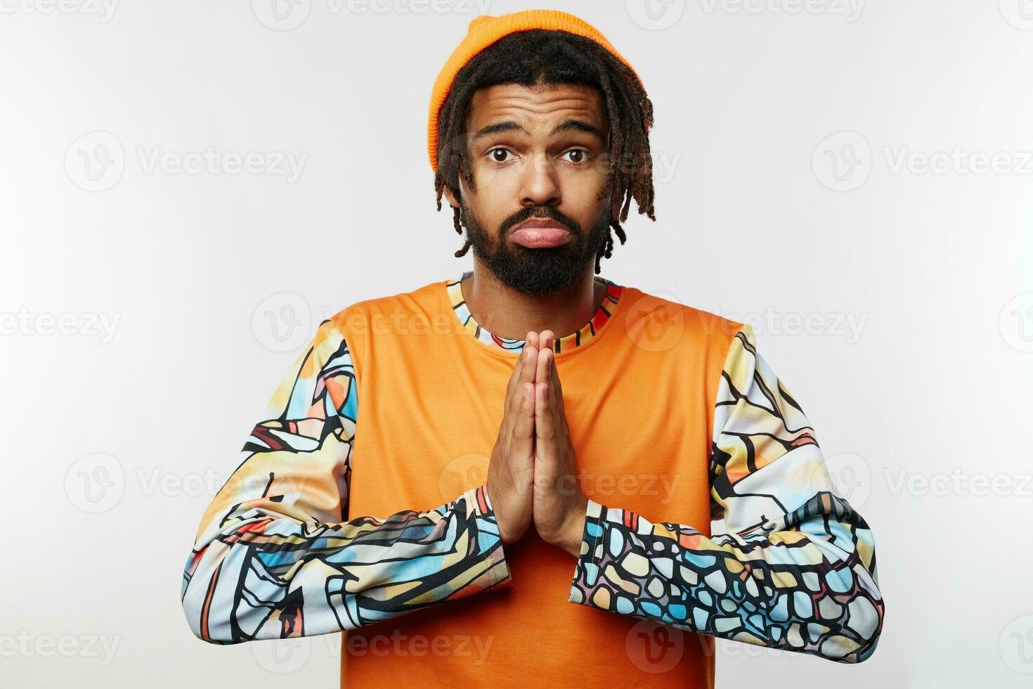 Confused young pretty bearded brunette guy with dreadlocks wrinkling forehead while looking at camera and folding raised hands, isolated over white background photo