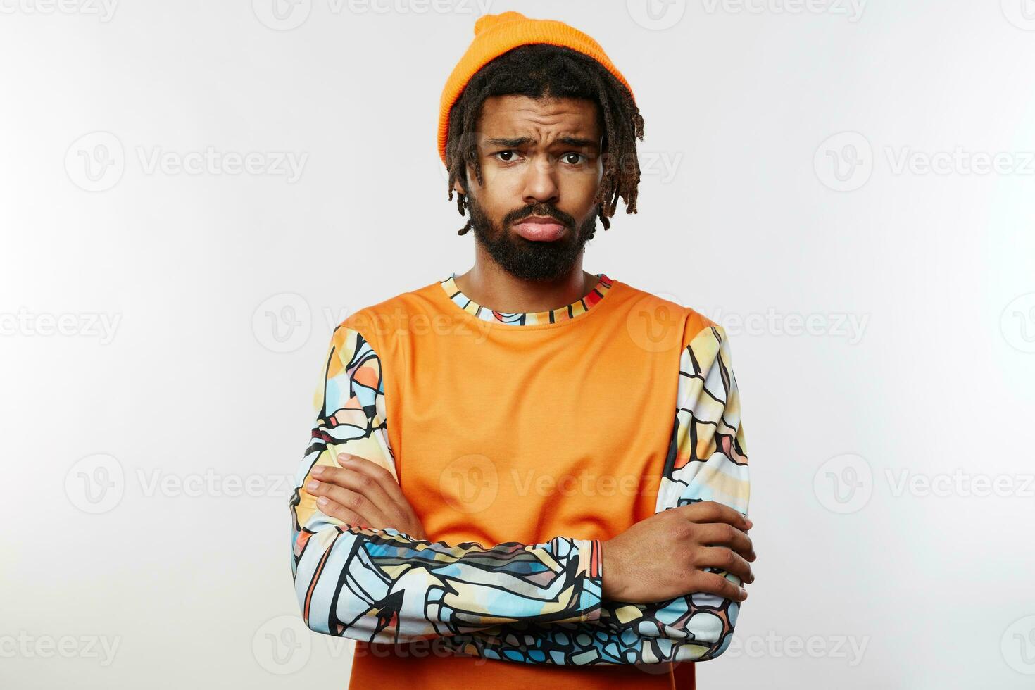 Unhappy young dark skinned brunette man crossing his arms while posing over white background, frowning his eyebrows while looking sadly at camera with folded lips photo