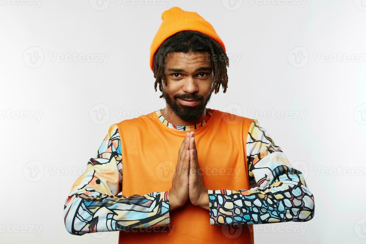 Glad young handsome dark skinned male with beard keeping raised palms together while looking positively at camera with light smile, isolated over white background photo