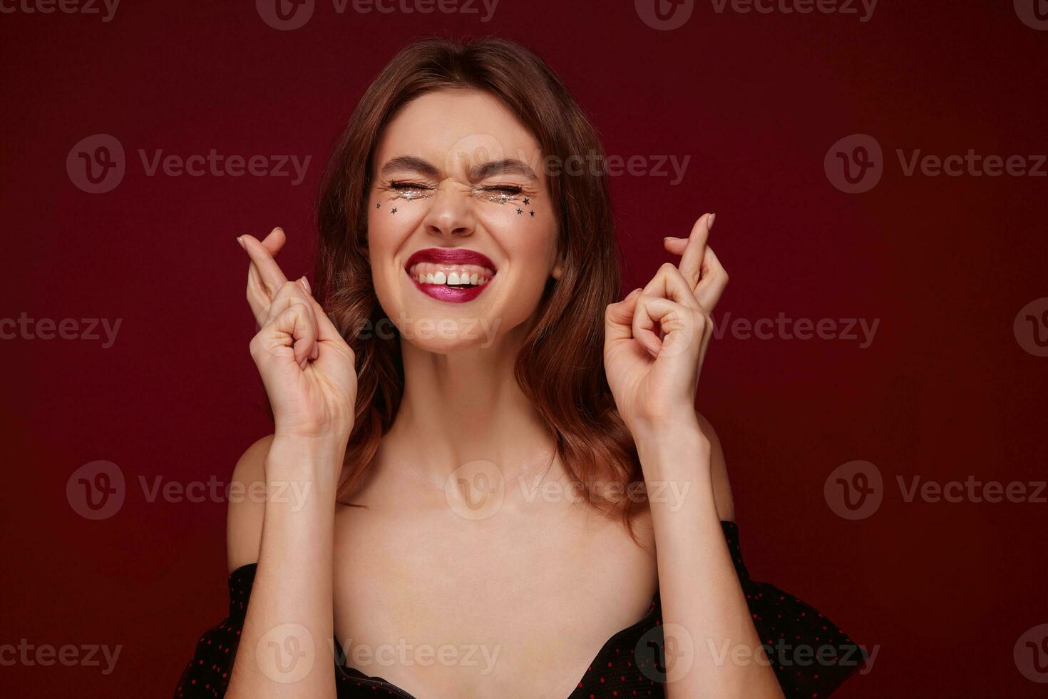 Hoping good looking young brunette woman keeping eyes closed while making wish, crossing fingers for good luck, wearing evening makeup and elegant clothes over claret background photo